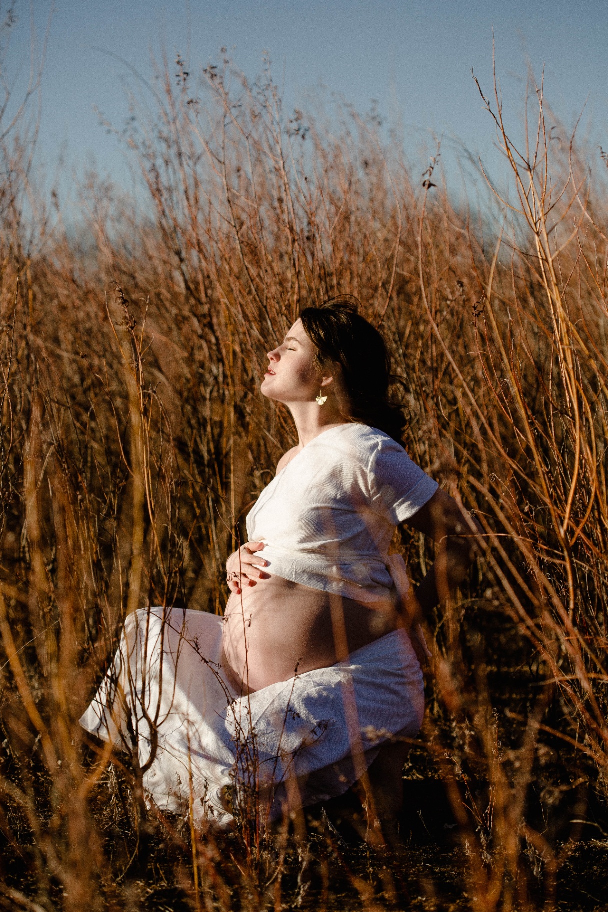 A woman in white kneeling in tall golden grasses, face lifted to the sun, hands cradling her belly